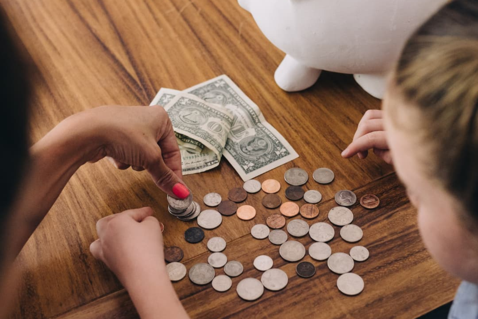 Niño y adulto contando monedas sobre una mesa junto a una alcancía blanca y billetes de dólar, representando enseñanza de educación financiera infantil y hábitos de ahorro desde casa.