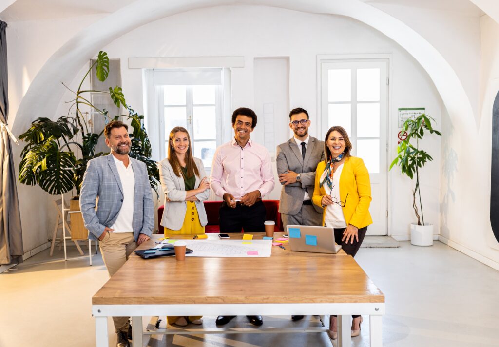 “Cinco profesionales reunidos alrededor de una mesa en una oficina moderna, colaborando sobre documentos y tecnología en un entorno luminoso con plantas y ventanas grandes.”