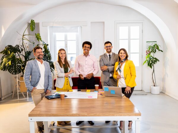 “Cinco profesionales reunidos alrededor de una mesa en una oficina moderna, colaborando sobre documentos y tecnología en un entorno luminoso con plantas y ventanas grandes.”